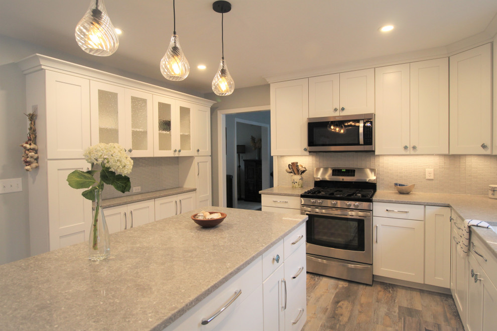 Photo of a mid-sized beach style l-shaped eat-in kitchen in Burlington with an undermount sink, shaker cabinets, white cabinets, quartz benchtops, grey splashback, glass tile splashback, stainless steel appliances, vinyl floors, with island, multi-coloured floor and grey benchtop.