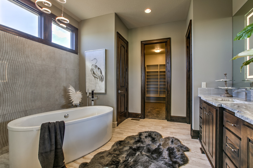 Photo of a transitional bathroom in Kansas City with raised-panel cabinets, brown cabinets, a freestanding tub, gray tile, subway tile, grey walls, an undermount sink, beige floor and grey benchtops.