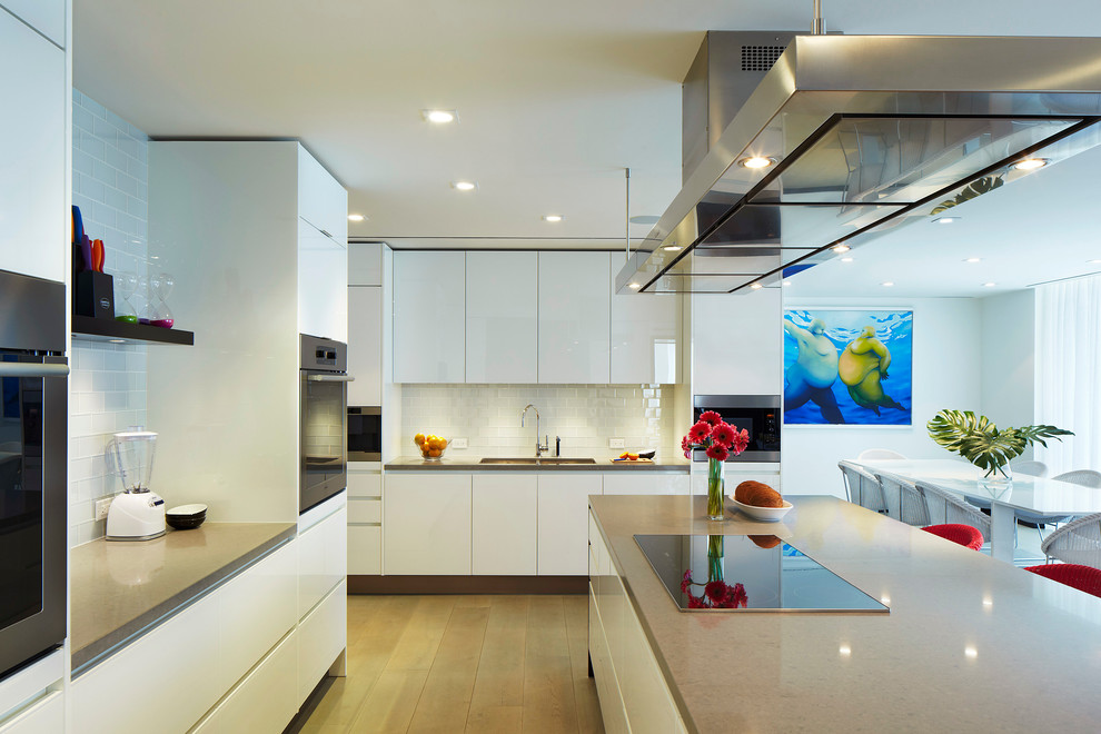 Photo of a large contemporary u-shaped eat-in kitchen in Miami with an undermount sink, flat-panel cabinets, white cabinets, quartz benchtops, white splashback, subway tile splashback, stainless steel appliances, light hardwood floors and with island.