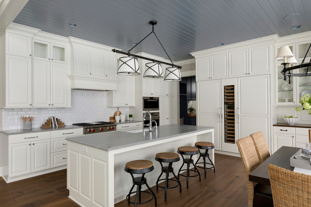 Photo of a traditional eat-in kitchen in Minneapolis with a farmhouse sink, raised-panel cabinets, white cabinets, white splashback, subway tile splashback, panelled appliances, with island, grey benchtop and medium hardwood floors.