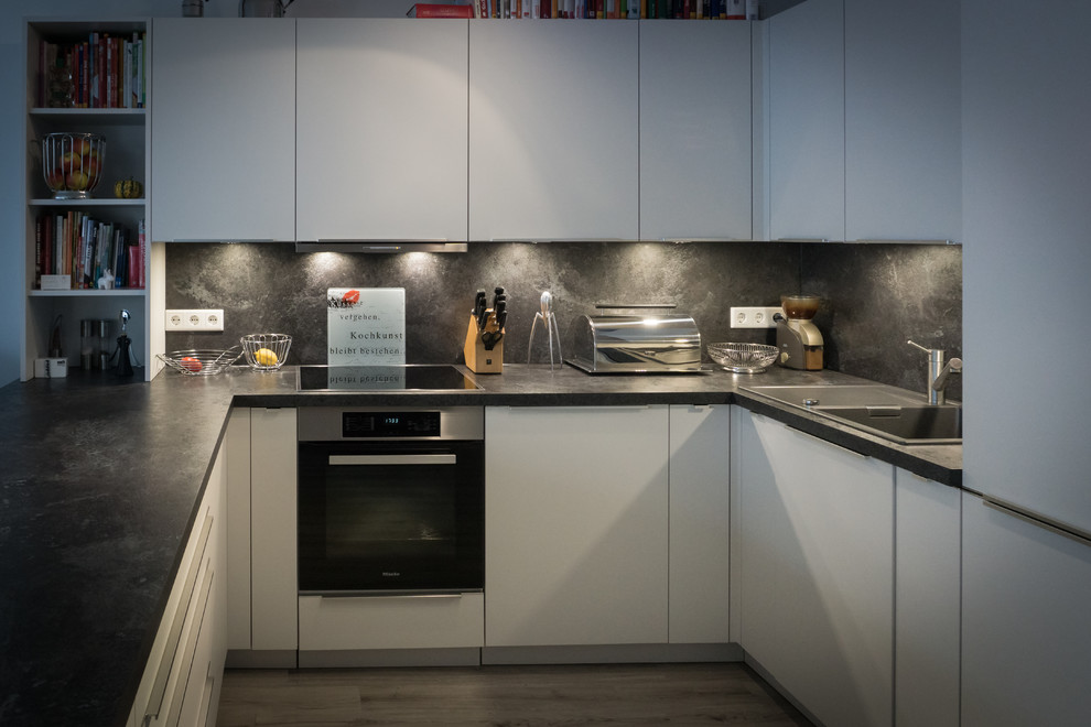 Design ideas for an expansive contemporary u-shaped open plan kitchen in Nuremberg with a drop-in sink, flat-panel cabinets, white cabinets, laminate benchtops, black splashback, timber splashback, black appliances, vinyl floors, a peninsula and black benchtop.