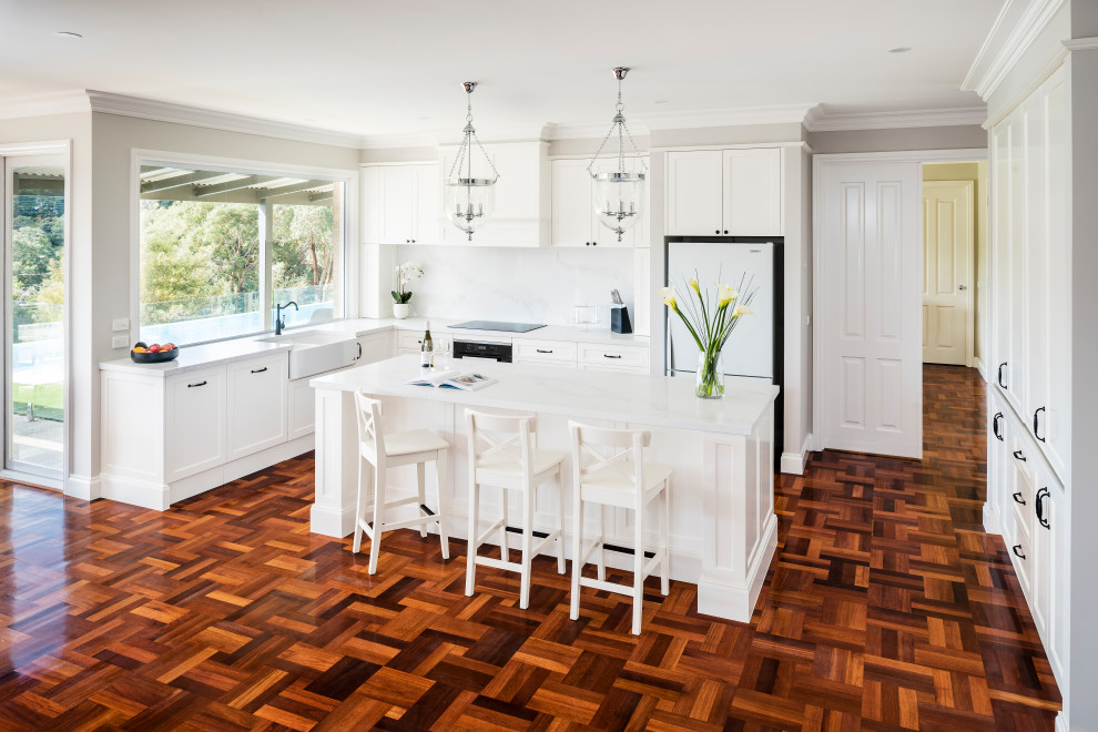 This is an example of a large traditional l-shaped eat-in kitchen in Melbourne with shaker cabinets, with island, a farmhouse sink, white cabinets, quartz benchtops, engineered quartz splashback, black appliances and dark hardwood floors.