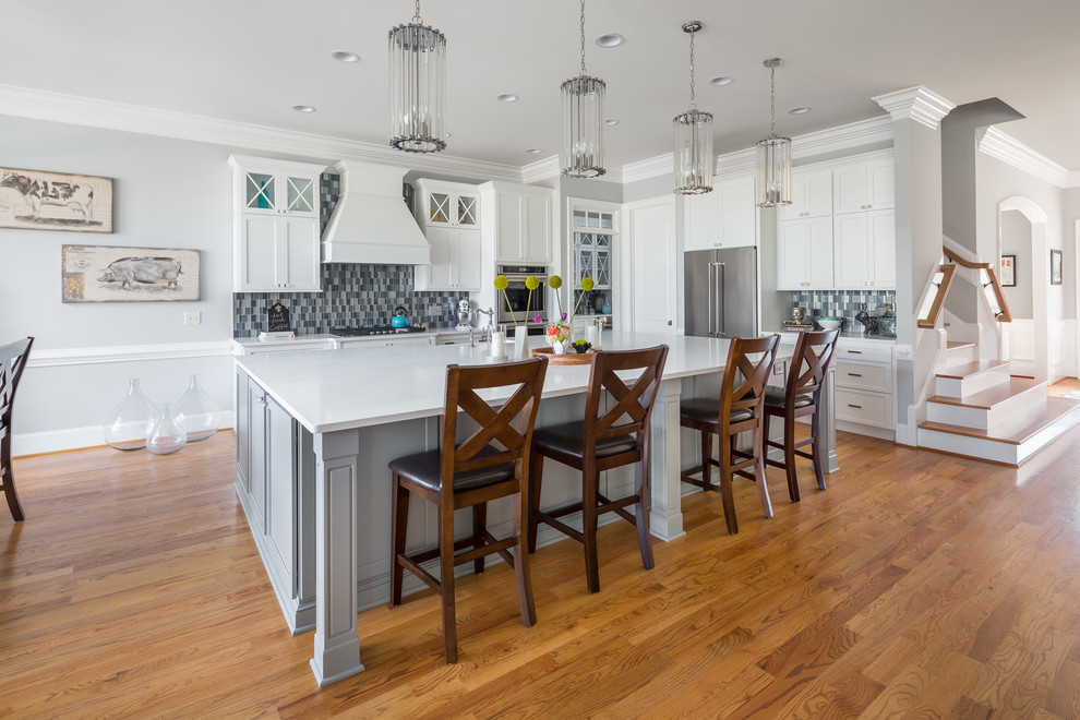 Example of a large transitional l-shaped medium tone wood floor open concept kitchen design in Raleigh with a farmhouse sink, flat-panel cabinets, white cabinets, quartz countertops, blue backsplash, glass tile backsplash, stainless steel appliances and an island