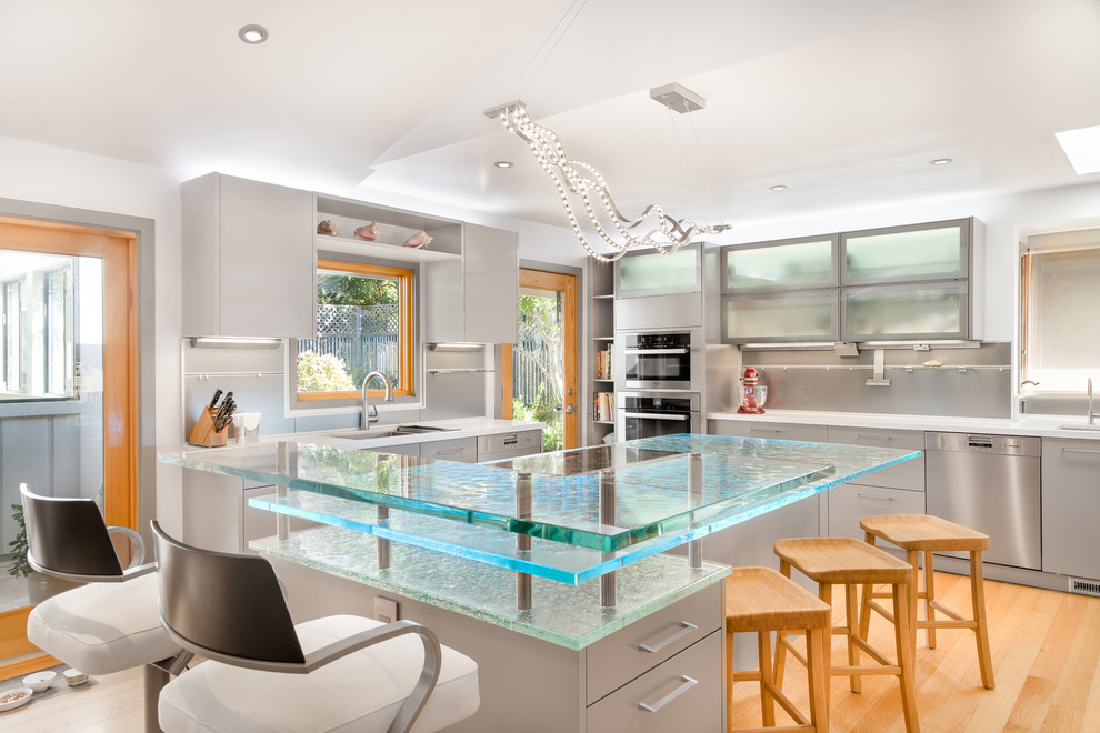 Photo of a large contemporary u-shaped kitchen in Los Angeles with flat-panel cabinets, glass benchtops, stainless steel appliances, light hardwood floors, an undermount sink, grey cabinets, grey splashback, with island and turquoise benchtop.