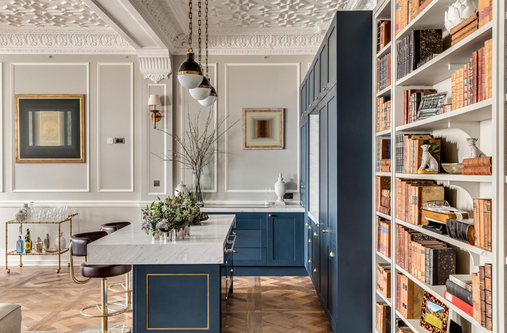 This is an example of an expansive traditional l-shaped home bar in London with an integrated sink, shaker cabinets, blue cabinets, marble benchtops, white splashback, marble splashback, medium hardwood floors, brown floor and white benchtop.
