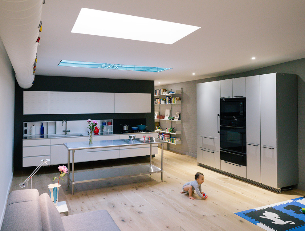 Inspiration for a contemporary open plan kitchen in New York with an integrated sink, flat-panel cabinets, stainless steel benchtops, white splashback, black appliances, light hardwood floors and with island.