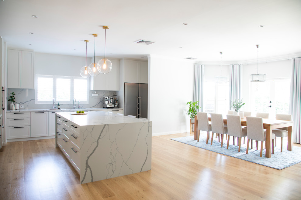 This is an example of an expansive transitional l-shaped open plan kitchen in Sydney with a double-bowl sink, shaker cabinets, white cabinets, quartz benchtops, white splashback, marble splashback, stainless steel appliances, light hardwood floors, with island, beige floor and white benchtop.