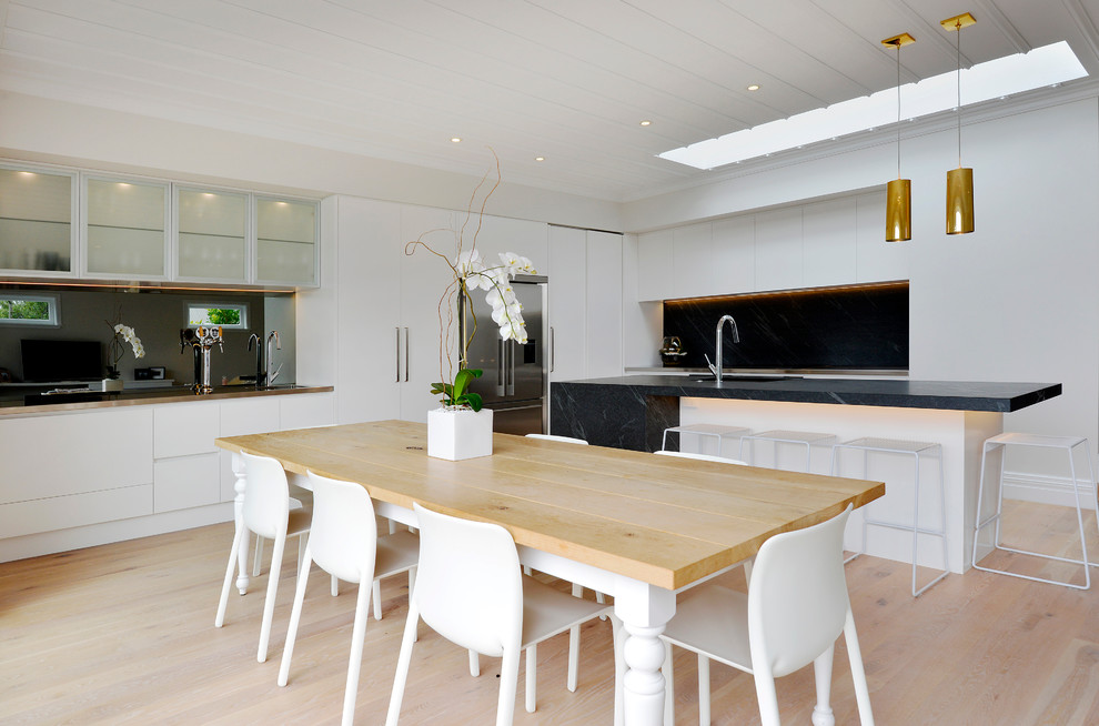 Photo of a mid-sized contemporary l-shaped eat-in kitchen in Auckland with an undermount sink, flat-panel cabinets, white cabinets, granite benchtops, black splashback, stone slab splashback, stainless steel appliances, light hardwood floors and with island.