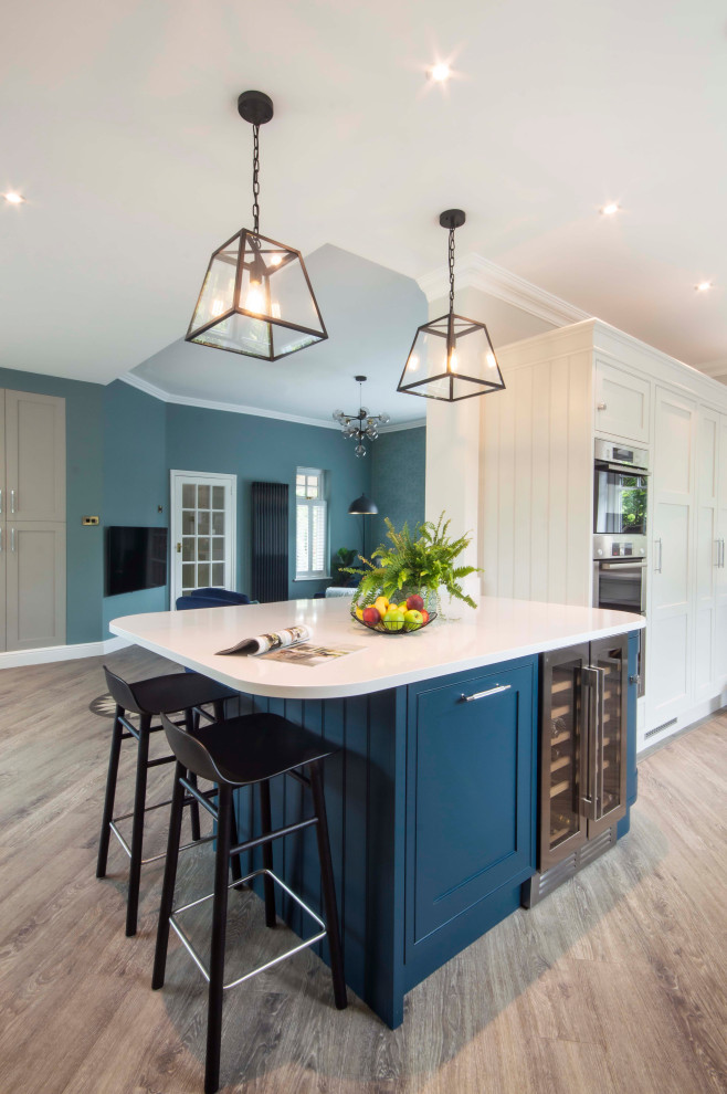 This is an example of a medium sized classic u-shaped open plan kitchen in Surrey with a belfast sink, shaker cabinets, blue cabinets, composite countertops, mirror splashback, integrated appliances, medium hardwood flooring, an island, brown floors, white worktops and a chimney breast.