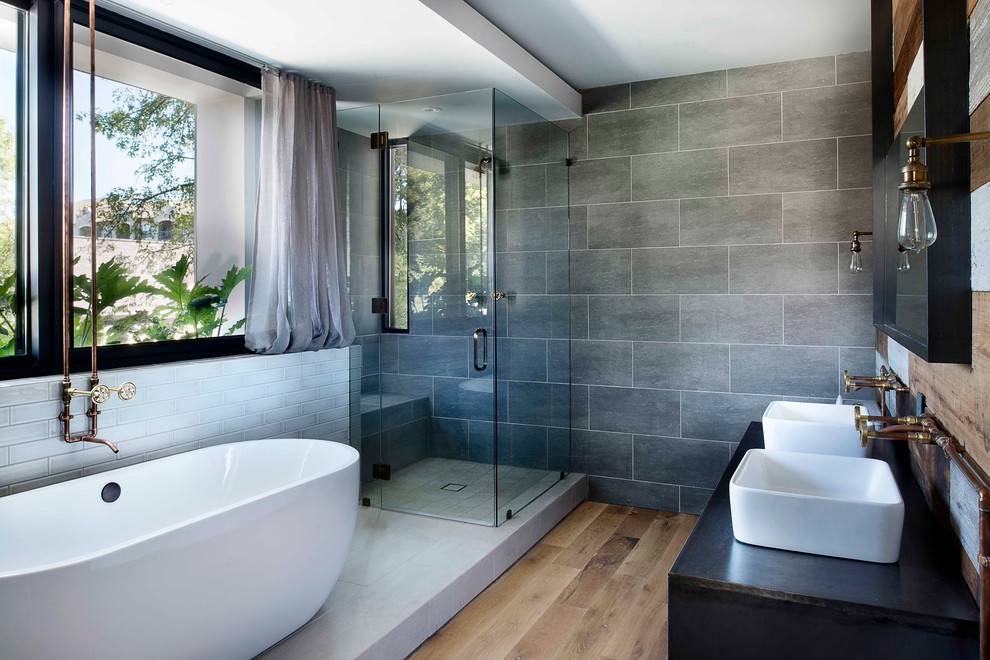 Photo of an industrial master bathroom in Atlanta with a freestanding tub, a curbless shower, gray tile, light hardwood floors, a vessel sink and a hinged shower door.