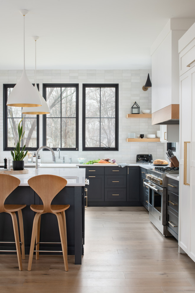 Example of a transitional l-shaped light wood floor kitchen design in DC Metro with a farmhouse sink, shaker cabinets, black cabinets, quartz countertops, white backsplash, ceramic backsplash, paneled appliances, an island and white countertops