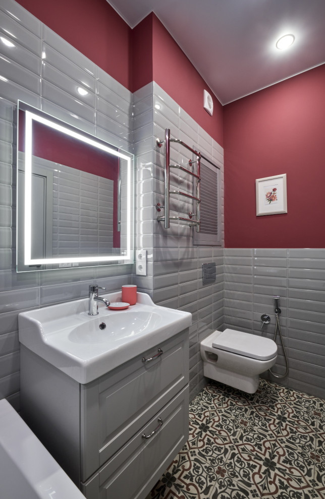 Mid-sized contemporary master bathroom in Moscow with raised-panel cabinets, grey cabinets, an undermount tub, a wall-mount toilet, gray tile, ceramic tile, pink walls, cement tiles, a wall-mount sink, multi-coloured floor, an enclosed toilet, a laundry, a single vanity and a floating vanity.