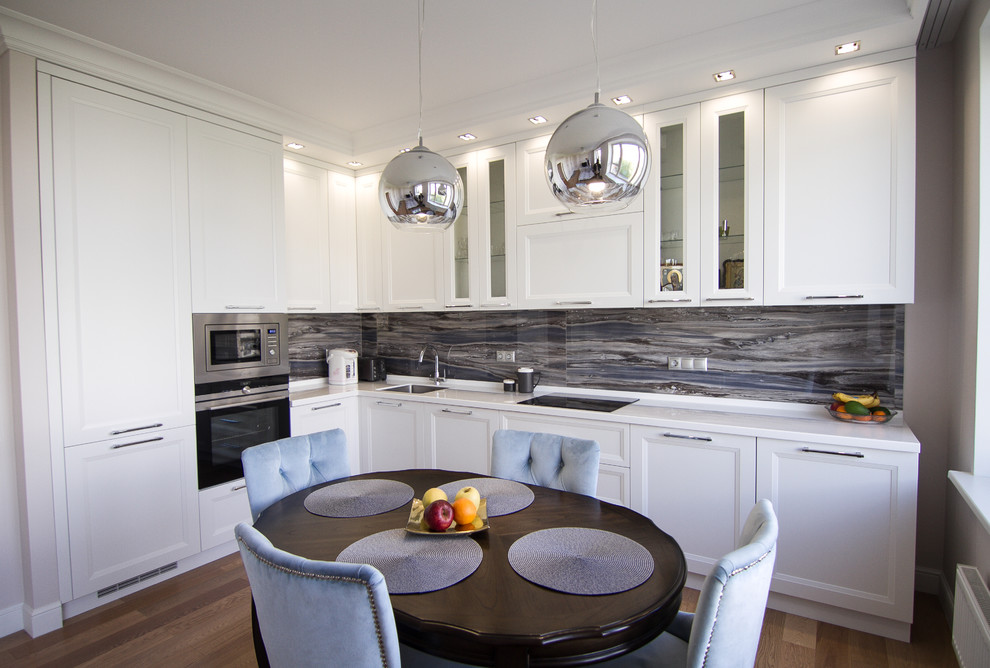 Photo of a mid-sized transitional l-shaped eat-in kitchen in Moscow with a drop-in sink, recessed-panel cabinets, white cabinets, solid surface benchtops, grey splashback, glass sheet splashback, stainless steel appliances, medium hardwood floors, no island, brown floor and white benchtop.