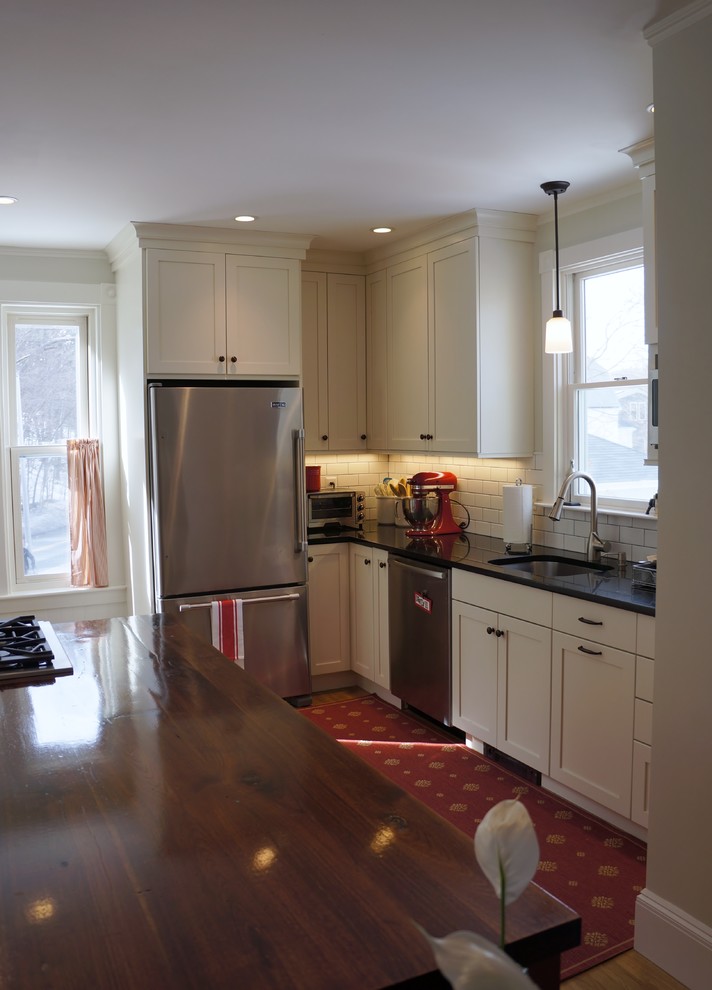 Small mountain style l-shaped light wood floor and beige floor enclosed kitchen photo in Boston with an undermount sink, recessed-panel cabinets, granite countertops, white backsplash, subway tile backsplash, stainless steel appliances, an island, white cabinets and black countertops