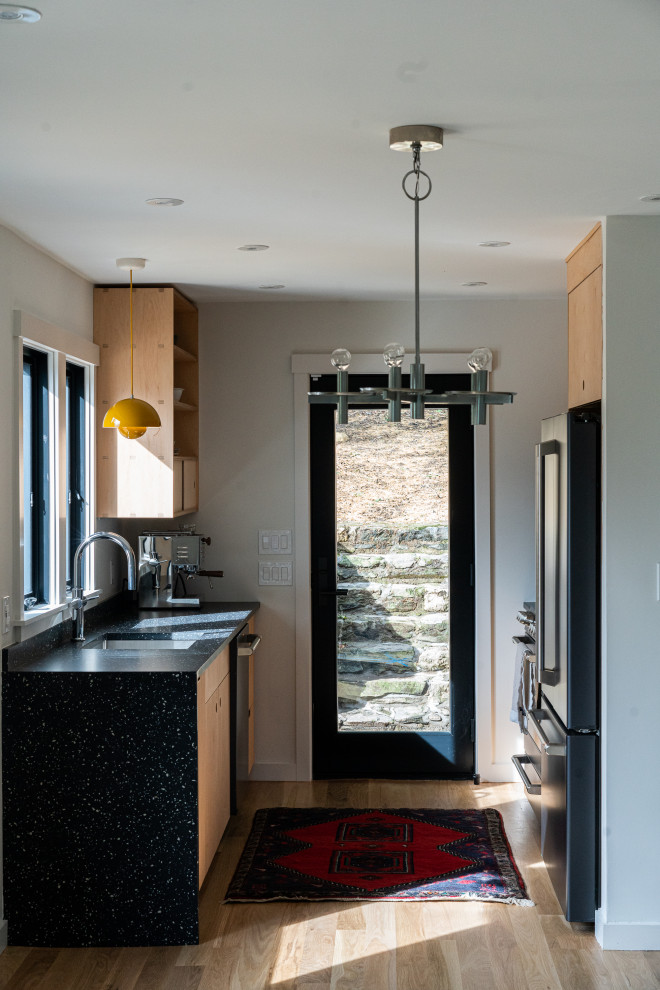 Cette image montre une petite cuisine parallèle minimaliste en bois clair avec un évier encastré, un placard à porte plane, un plan de travail en terrazzo, une crédence noire, un électroménager noir, parquet clair et plan de travail noir.