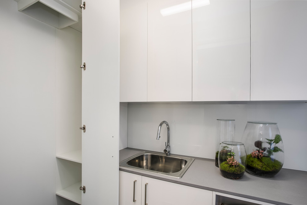 Mid-sized contemporary single-wall laundry room in Sydney with a drop-in sink, white cabinets, quartz benchtops, white walls, ceramic floors, grey floor and grey benchtop.