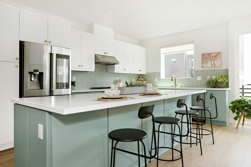 Contemporary l-shaped open plan kitchen in Los Angeles with an undermount sink, flat-panel cabinets, grey splashback, stainless steel appliances, light hardwood floors, with island, brown floor, white benchtop and green cabinets.