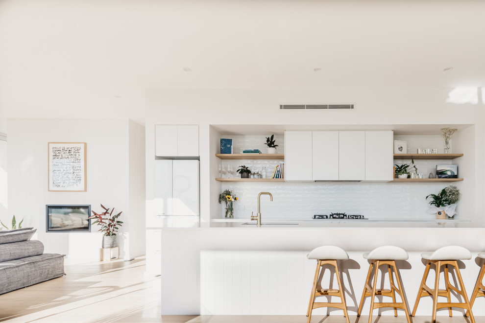 Photo of a contemporary galley kitchen in Sydney with white splashback, subway tile splashback, with island, flat-panel cabinets, white cabinets, white appliances, light hardwood floors, beige floor and white benchtop.