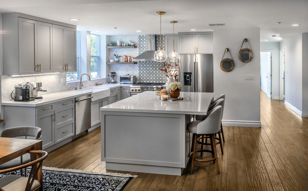 Mid-sized country l-shaped eat-in kitchen in Los Angeles with a farmhouse sink, shaker cabinets, grey cabinets, quartz benchtops, white splashback, marble splashback, stainless steel appliances, medium hardwood floors, with island and brown floor.