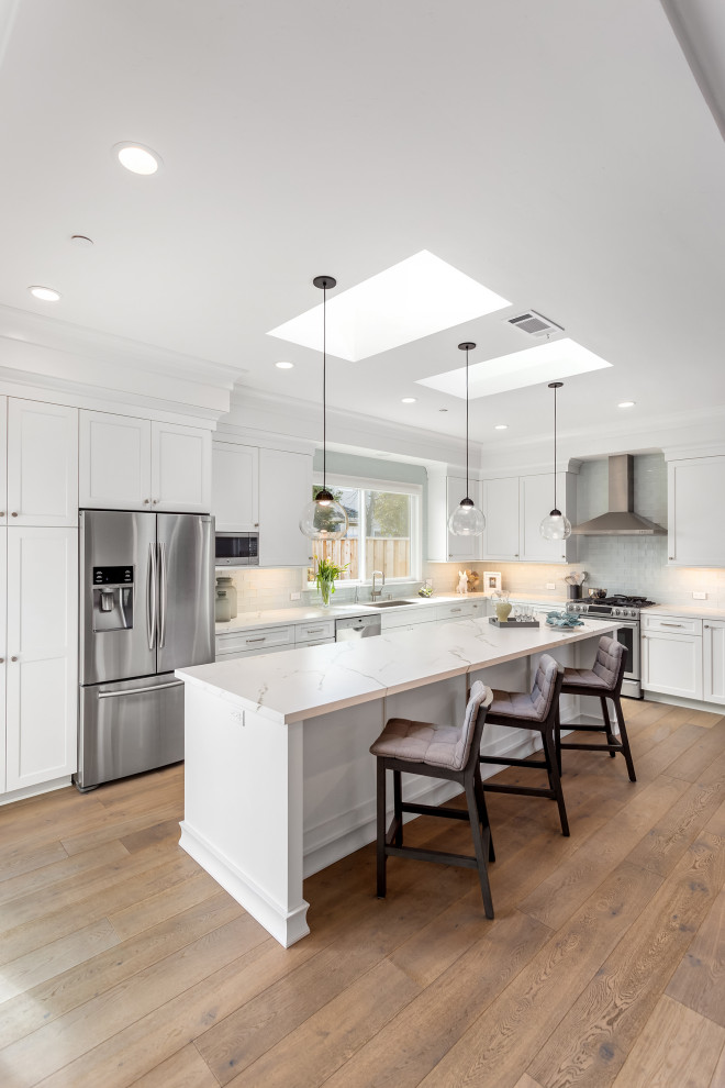 Design ideas for a large transitional l-shaped open plan kitchen in San Francisco with an undermount sink, shaker cabinets, white cabinets, quartz benchtops, white splashback, ceramic splashback, stainless steel appliances, light hardwood floors, with island and white benchtop.