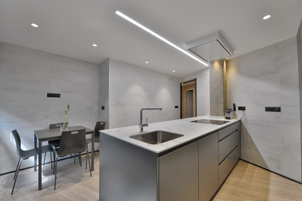 This is an example of a mid-sized industrial galley open plan kitchen in Madrid with an undermount sink, flat-panel cabinets, grey cabinets, limestone benchtops, stainless steel appliances, laminate floors, a peninsula, beige floor and white benchtop.