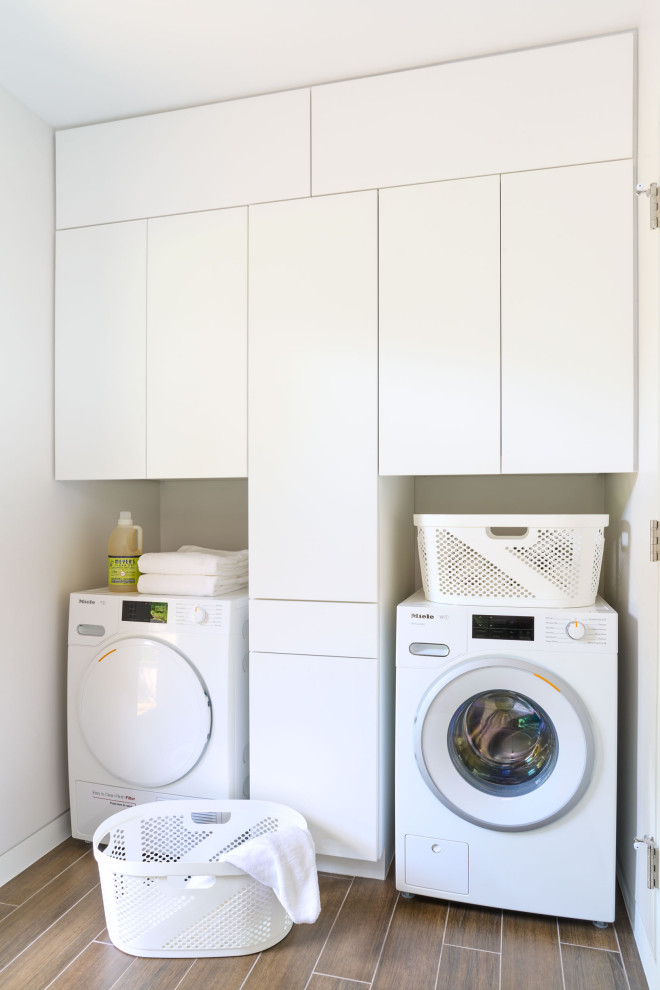 Foto på en liten funkis linjär tvättstuga enbart för tvätt, med släta luckor, vita skåp, vita väggar, klinkergolv i keramik, en tvättmaskin och torktumlare bredvid varandra och brunt golv