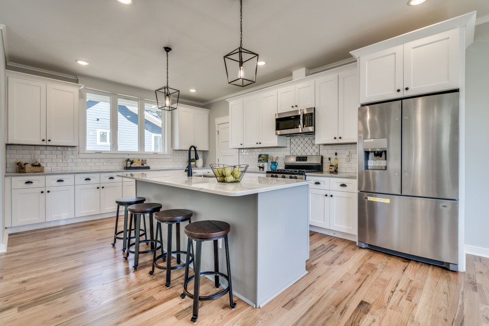 This is an example of a large transitional l-shaped eat-in kitchen in Raleigh with a single-bowl sink, shaker cabinets, white cabinets, quartzite benchtops, subway tile splashback, stainless steel appliances, light hardwood floors, with island, white benchtop, white splashback and beige floor.