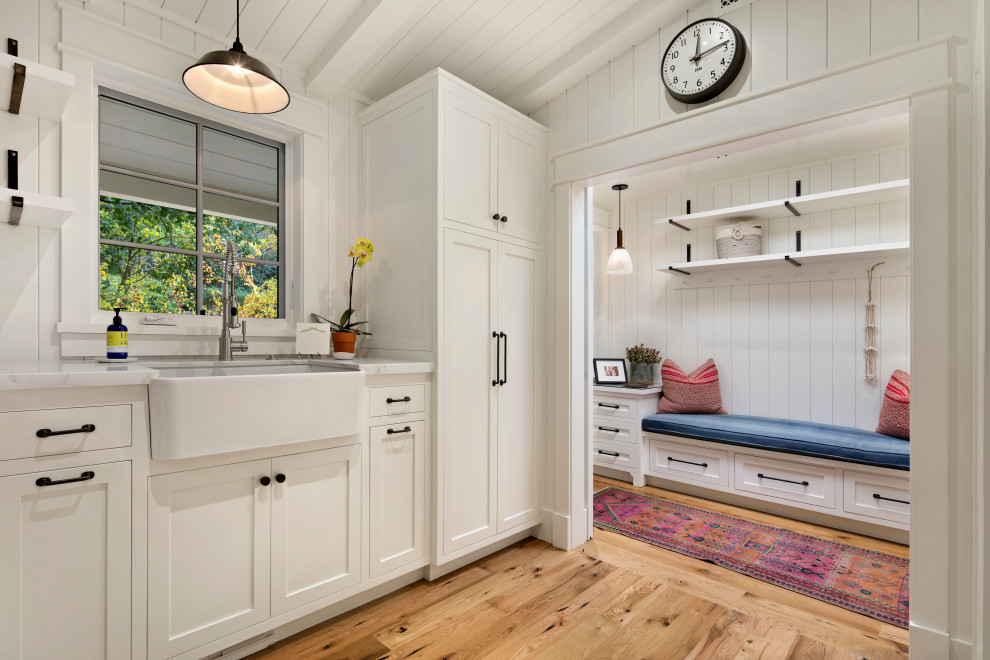 Large modern u-shaped dedicated laundry room in San Francisco with a farmhouse sink, shaker cabinets, white cabinets, marble benchtops, white splashback, timber splashback, white walls, light hardwood floors, a side-by-side washer and dryer, white benchtop, vaulted and planked wall panelling.