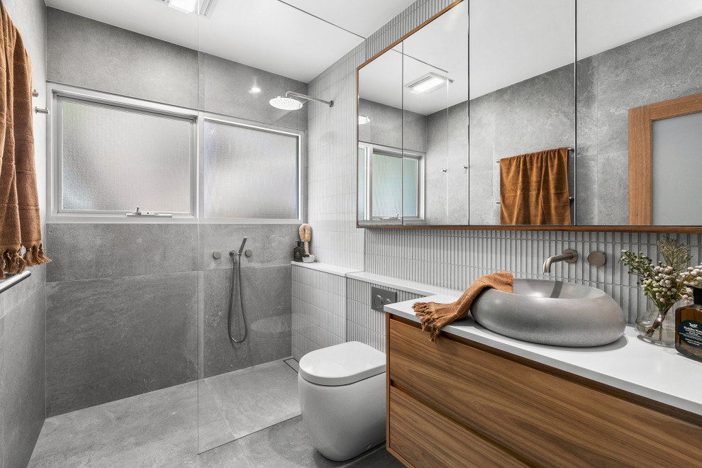 Photo of a medium sized contemporary ensuite bathroom in Sydney with freestanding cabinets, medium wood cabinets, a walk-in shower, a one-piece toilet, grey tiles, porcelain tiles, grey walls, porcelain flooring, a vessel sink, solid surface worktops, grey floors, an open shower, white worktops, a single sink and a built in vanity unit.
