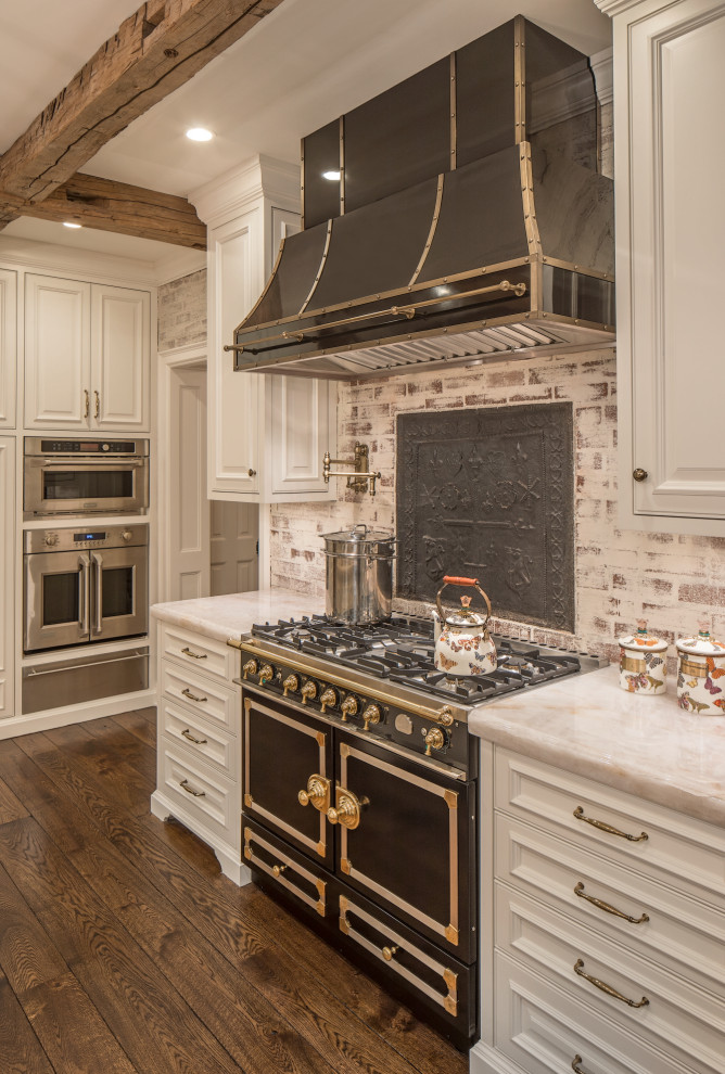 Inspiration for a large traditional l-shaped kitchen in Omaha with a farmhouse sink, raised-panel cabinets, white cabinets, granite benchtops, white splashback, brick splashback, stainless steel appliances, medium hardwood floors, with island, brown floor and beige benchtop.
