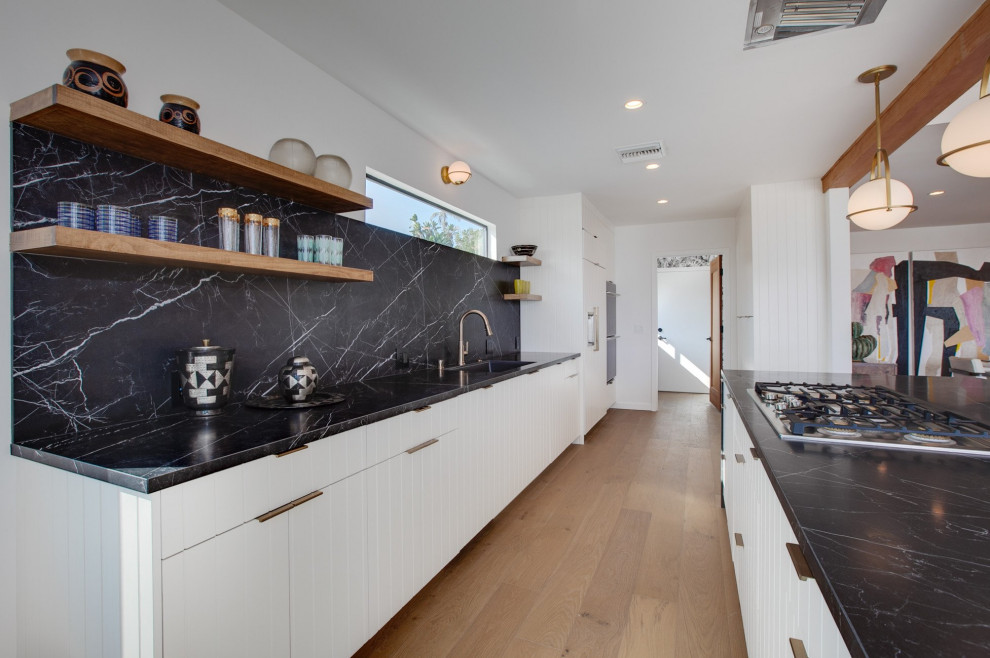 Photo of a midcentury galley kitchen in Los Angeles with an undermount sink, flat-panel cabinets, white cabinets, black splashback, stone slab splashback, panelled appliances, medium hardwood floors, with island, brown floor and black benchtop.