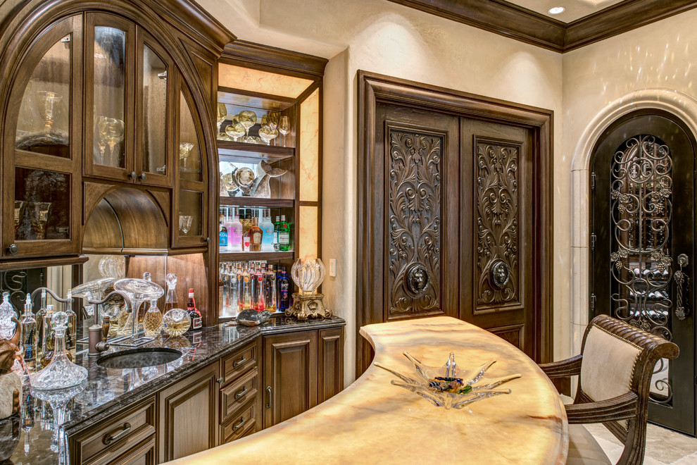 This is an example of a mediterranean u-shaped seated home bar with a drop-in sink, raised-panel cabinets, dark wood cabinets, onyx benchtops, mirror splashback, travertine floors, beige floor and beige benchtop.
