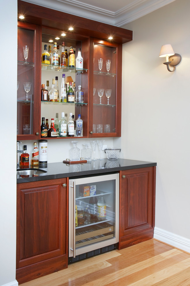 This is an example of a traditional single-wall wet bar in Perth with an undermount sink, medium wood cabinets, quartz benchtops, black benchtop, light hardwood floors, beige floor and raised-panel cabinets.