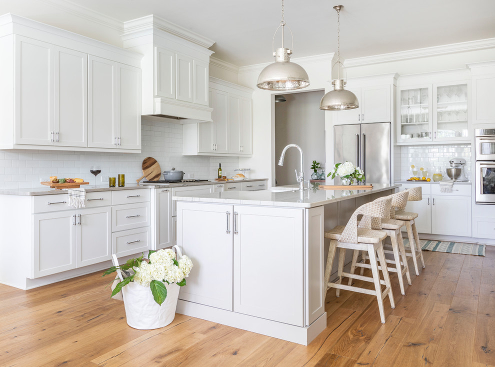 Photo of a beach style l-shaped kitchen in Jacksonville with a farmhouse sink, shaker cabinets, white cabinets, white splashback, subway tile splashback, stainless steel appliances, light hardwood floors, with island, beige floor and beige benchtop.