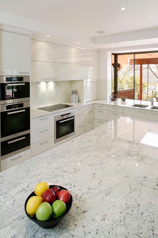 This is an example of an u-shaped eat-in kitchen in Sydney with a drop-in sink, flat-panel cabinets, white cabinets, solid surface benchtops, white splashback, glass sheet splashback, stainless steel appliances, ceramic floors and with island.