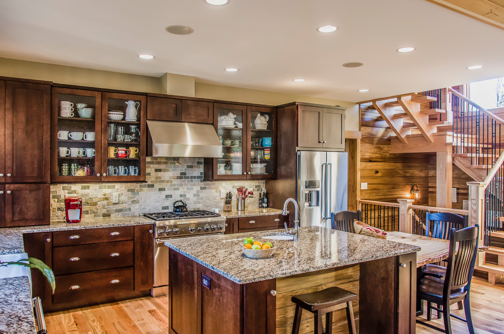 This is an example of a large country l-shaped eat-in kitchen in Other with shaker cabinets, dark wood cabinets, granite benchtops, beige splashback, stone tile splashback, stainless steel appliances, with island, a farmhouse sink, light hardwood floors, beige floor and beige benchtop.