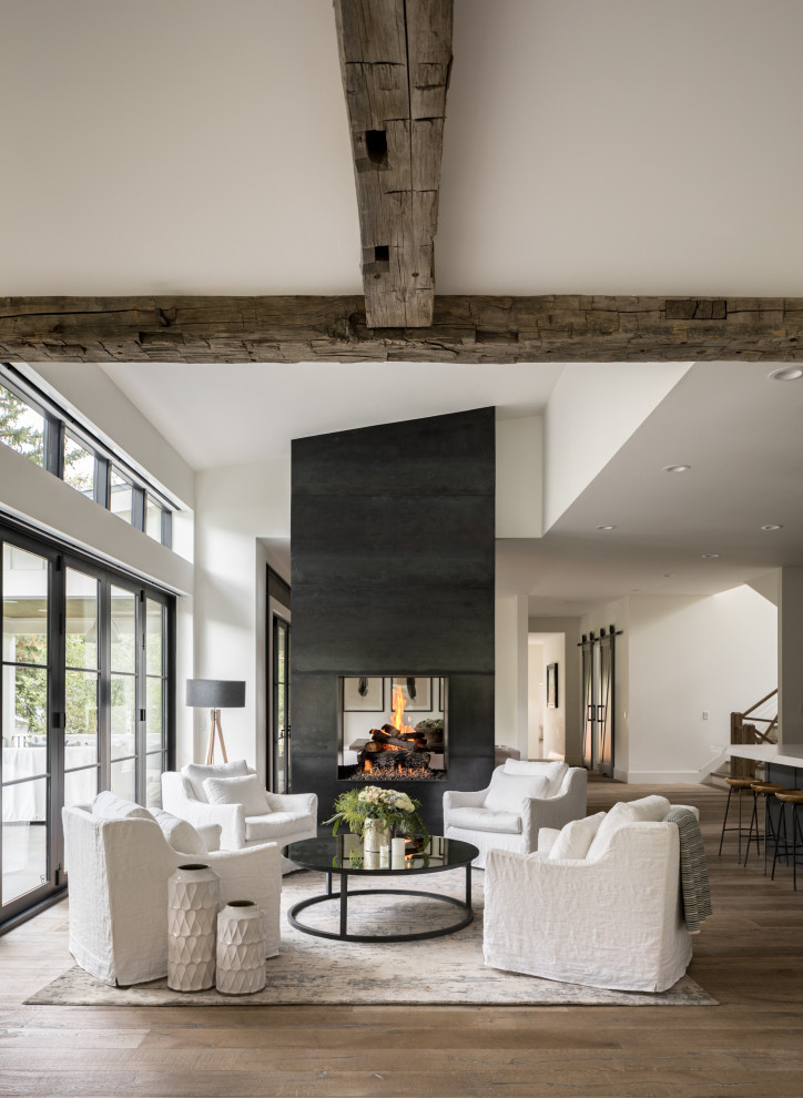 This is an example of a country open concept living room in Denver with white walls, medium hardwood floors, a two-sided fireplace, a metal fireplace surround, grey floor and exposed beam.