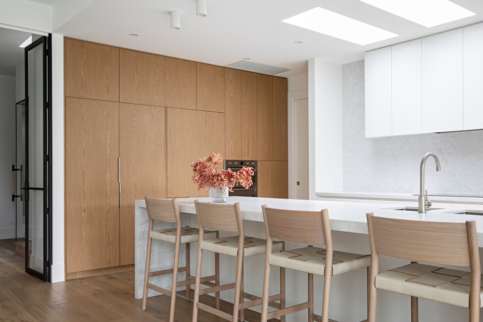 Photo of a contemporary l-shaped kitchen in Melbourne with an undermount sink, flat-panel cabinets, medium wood cabinets, white splashback, stone slab splashback, stainless steel appliances, medium hardwood floors, with island, brown floor and white benchtop.