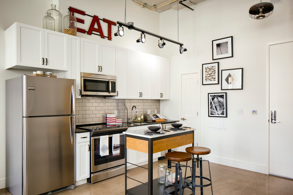 Small industrial single-wall eat-in kitchen in DC Metro with white cabinets, beige splashback, stainless steel appliances, an undermount sink, recessed-panel cabinets, quartz benchtops, subway tile splashback, linoleum floors, with island, brown floor and black benchtop.