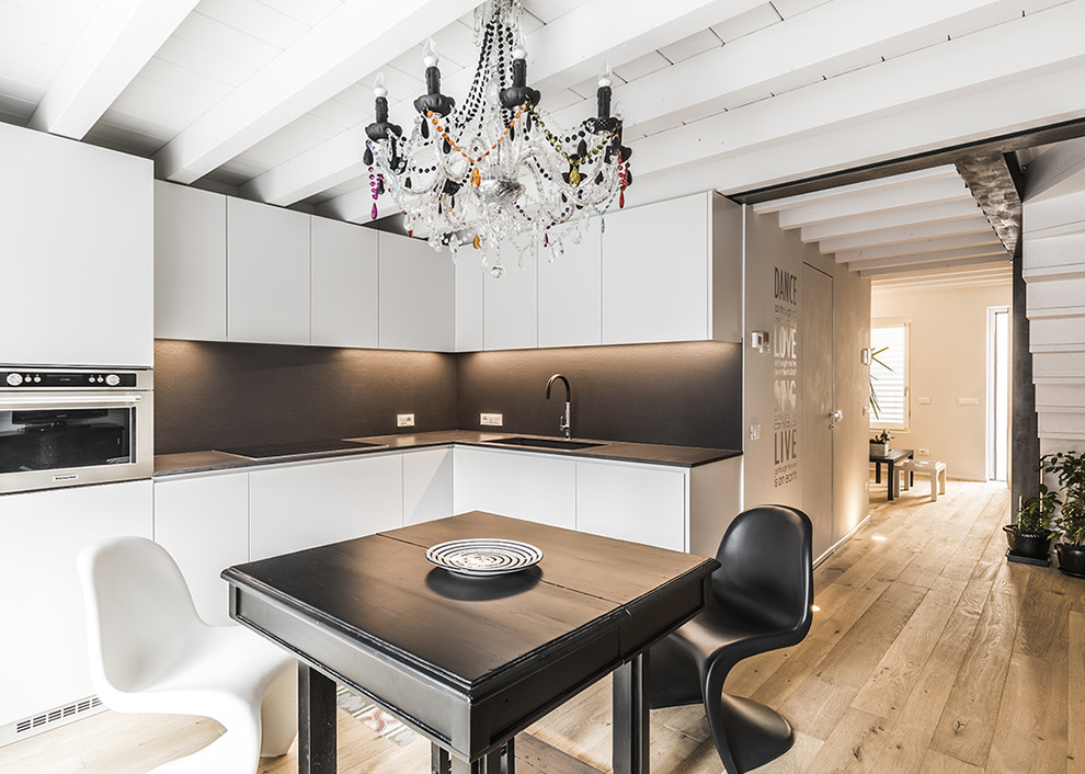 Mid-sized contemporary l-shaped eat-in kitchen in Other with an undermount sink, flat-panel cabinets, white cabinets, black splashback, stainless steel appliances, medium hardwood floors, no island, brown floor and black benchtop.