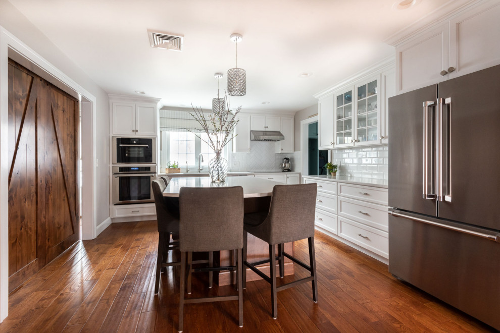 Large transitional l-shaped medium tone wood floor and brown floor eat-in kitchen photo in Boston with an undermount sink, shaker cabinets, white cabinets, quartz countertops, white backsplash, subway tile backsplash, stainless steel appliances, an island and white countertops