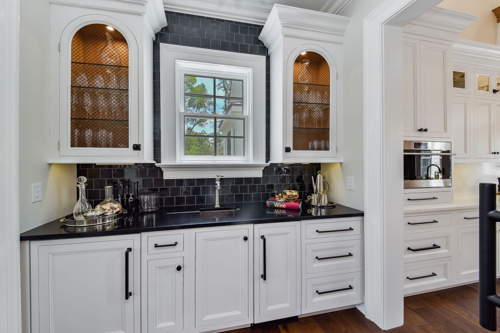 This is an example of a mid-sized traditional single-wall wet bar in Chicago with an undermount sink, white cabinets, quartz benchtops, black splashback, ceramic splashback, dark hardwood floors, black benchtop, recessed-panel cabinets and brown floor.