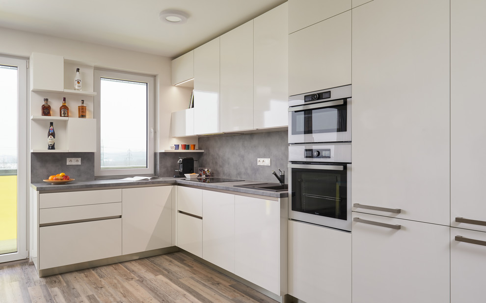 Photo of a mid-sized contemporary l-shaped eat-in kitchen in Other with a drop-in sink, flat-panel cabinets, white cabinets, laminate benchtops, grey splashback, white appliances, plywood floors and no island.