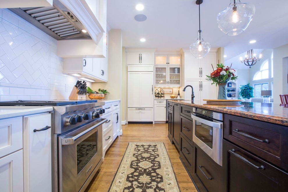 Large transitional l-shaped light wood floor and brown floor enclosed kitchen photo with a farmhouse sink, shaker cabinets, white cabinets, solid surface countertops, white backsplash, porcelain backsplash, stainless steel appliances and an island