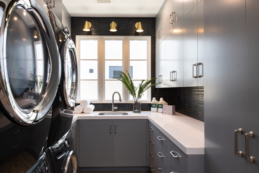 This is an example of a mid-sized transitional u-shaped dedicated laundry room in Orange County with an undermount sink, flat-panel cabinets, grey cabinets, quartz benchtops, black splashback, ceramic splashback, black walls, vinyl floors, a stacked washer and dryer, grey floor and white benchtop.