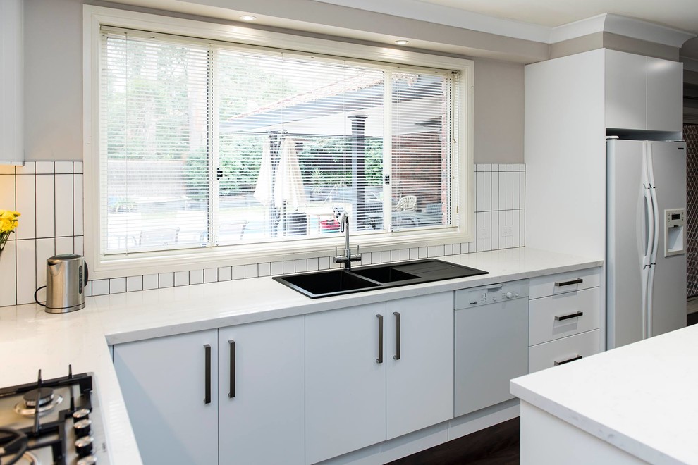 Photo of a large modern l-shaped eat-in kitchen in Sydney with a drop-in sink, flat-panel cabinets, white cabinets, quartz benchtops, white splashback, ceramic splashback, white appliances, dark hardwood floors, with island and white benchtop.