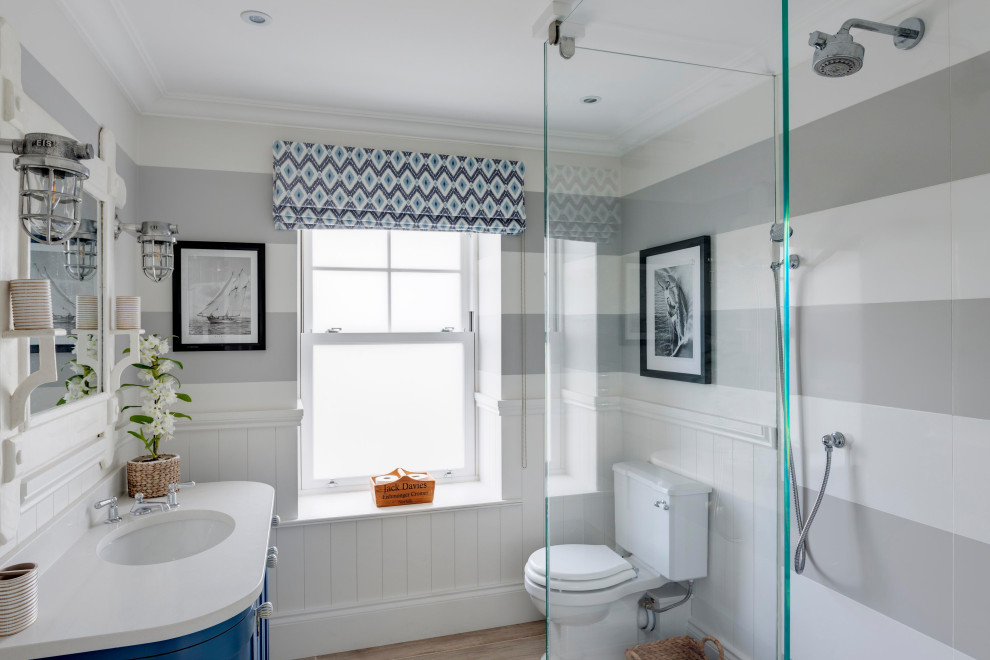 This is an example of a mid-sized beach style master bathroom in Devon with flat-panel cabinets, distressed cabinets, a curbless shower, a wall-mount toilet, blue tile, blue walls, ceramic floors, an integrated sink, onyx benchtops, grey floor, an open shower, brown benchtops, a niche, a double vanity, a freestanding vanity, recessed and wallpaper.