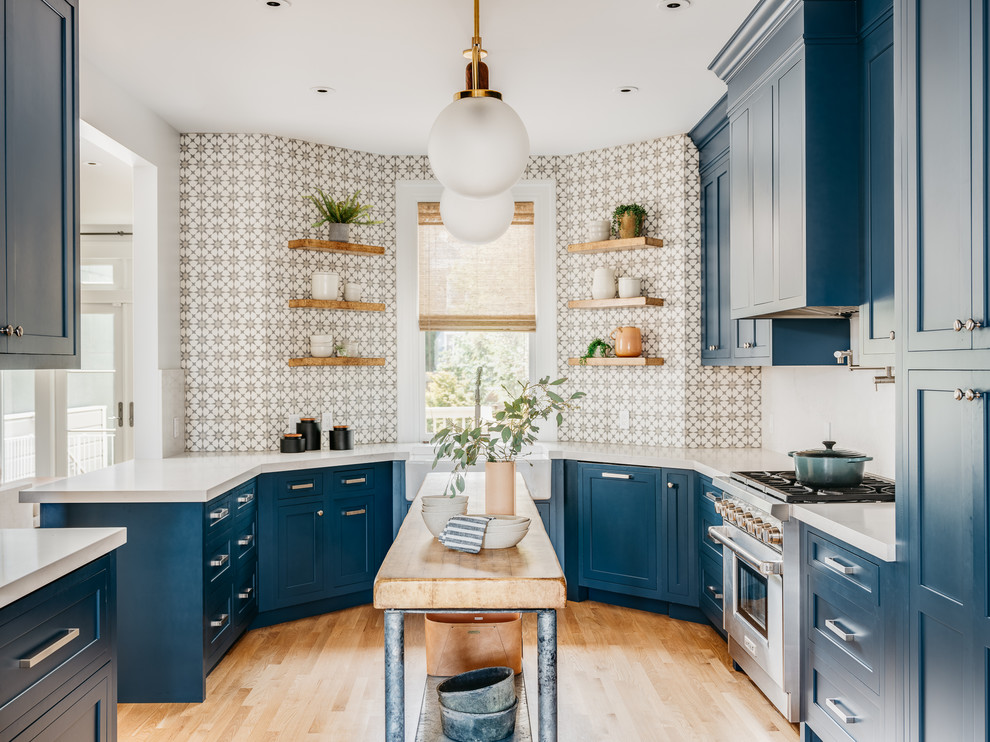 This is an example of a scandinavian u-shaped kitchen in San Francisco with a farmhouse sink, shaker cabinets, blue cabinets, multi-coloured splashback, stainless steel appliances, light hardwood floors, with island, white benchtop and beige floor.