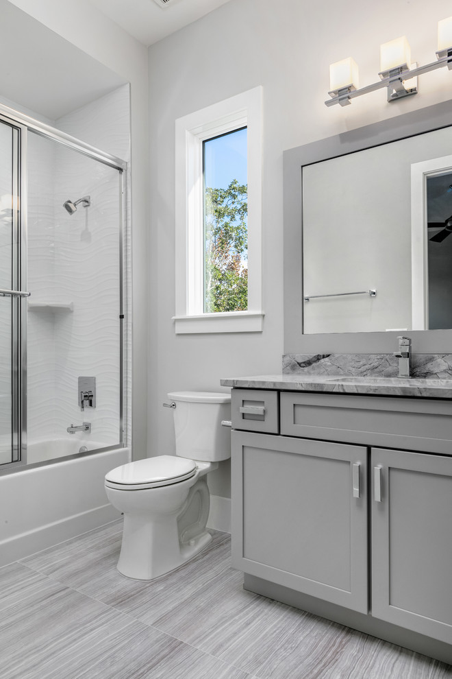 This is an example of a mid-sized transitional 3/4 bathroom in Orlando with shaker cabinets, grey cabinets, an alcove tub, a shower/bathtub combo, a two-piece toilet, white tile, grey walls, porcelain floors, an undermount sink, marble benchtops, grey floor, a sliding shower screen and grey benchtops.
