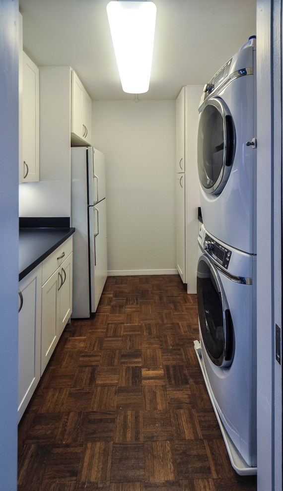 Inspiration for a mid-sized modern galley utility room in Baltimore with a double-bowl sink, recessed-panel cabinets, white cabinets, laminate benchtops, white walls, medium hardwood floors, a stacked washer and dryer, grey floor and black benchtop.